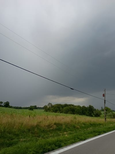 The image depicts an outdoor scene with a stormy sky looming overhead. A power line stretches diagonally across the frame, supported by a wooden pole with an orange electrical box attached. To the right of the power line, a rural landscape unfolds, featuring a grassy field and a line of trees. The sky appears overcast and dark, suggesting the presence of a storm. The lighting conditions are dim, contributing to a somewhat somber mood. There is no visible text in the image. The scene is characteristic of a rural area, potentially in Shelbyville, United States, given the provided location context. The composition, with the power line bisecting the sky and landscape, creates a sense of dramatic tension, perhaps amplified by the approaching storm.