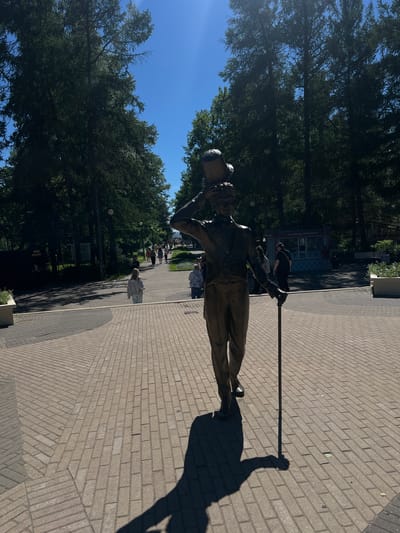 The image depicts a bronze statue of a man in a top hat and suit, holding a cane and balancing a bucket on his head. The statue is positioned on a brick-paved walkway in a park, with lush green trees lining the path and a clear blue sky overhead. Several people are visible in the background, walking along the path and near small kiosks. One kiosk has text in Russian that appears to advertise ice cream. The scene suggests a sunny day in a public recreational area, likely a park in Zelenogorsk, Russia, given the presence of Russian text and the typical appearance of such parks in the region. The statue casts a long shadow on the patterned brickwork.