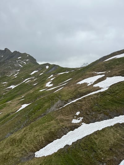 The image depicts a cloudy day in the mountainous region of Hasliberg Wasserwendi, Switzerland. The foreground is dominated by a steep, grassy slope dotted with patches of melting snow. The terrain is uneven, with visible rock formations and a narrow path winding through the vegetation. In the mid-ground, the slope continues, leading towards a ridgeline that stretches across the frame. More snow patches are scattered across the landscape, suggesting the remnants of a recent snowfall or the persistent presence of snow at higher altitudes. Further in the distance, the peaks of other mountains are visible, shrouded in a light mist or fog. A wooden fence runs along the top of one of the ridges, indicating a boundary or a walking trail. The sky is overcast with heavy, gray clouds, contributing to a somber and cool atmosphere. There are no people or animals visible in the image.