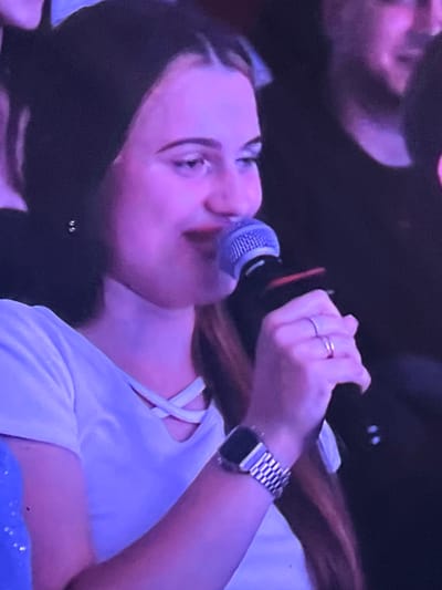 A young woman with dark hair and light skin, wearing a white t-shirt and a watch on her left wrist, holds a microphone to her mouth. Her eyes are directed to her right, and she has red lipstick on. She is wearing two rings on her right hand. The scene is dimly lit with purple and pink lighting. This image was taken in the city of Genemuiden, Netherlands.