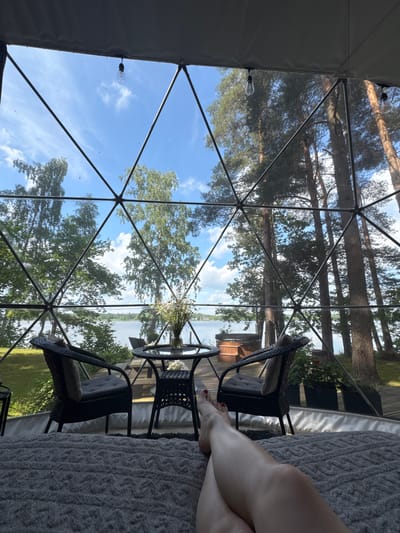 The image depicts a geodesic dome structure offering a view of a lake and surrounding trees. Inside the dome, a person's legs are visible in the foreground, resting on a gray rug. A glass table with chairs is set up outside the dome, along with a hot tub visible in the background. The scene is set in Priedesmuiža, Latvia.