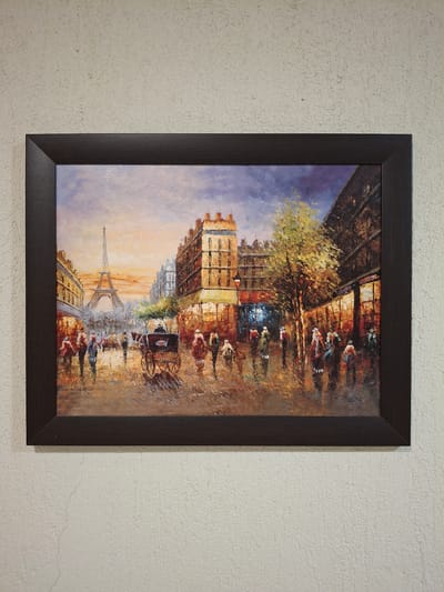 The image is a framed painting of a Parisian street scene featuring the Eiffel Tower in the distance. The painting depicts a street with buildings on either side, trees, and people walking. A horse-drawn carriage is also visible. The painting is in a dark brown frame and is hanging on a textured, off-white wall. The location is not in Krumovgrad, Bulgaria.
