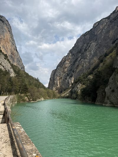 The image displays a deep natural gorge located in the vicinity of Fermignano, Italy. A broad expanse of calm, turquoise-green water occupies the central and lower portions of the frame. Flanking the water on both sides are towering, sheer rock cliffs, predominantly grey and beige in color, with visible vertical strata and fractured surfaces. The lower slopes of both cliffs are sparsely vegetated with deciduous and evergreen trees and shrubs. In the foreground, along the left side, a weathered concrete barrier with a dark, corroded metal handrail extends horizontally, indicating a pathway or road running alongside the water. The sky above is partially overcast with scattered white and grey cumulus clouds, allowing intermittent patches of brighter sky to be visible. No human figures, objects of human origin beyond the barrier, or actions are depicted in the scene.