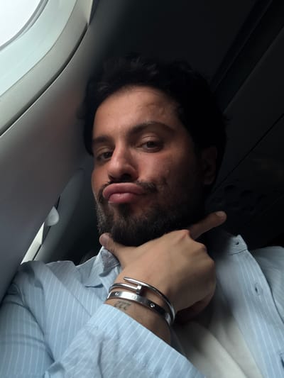A man with dark hair and a beard is resting his head against what appears to be the wall of an airplane cabin. He is wearing a light blue, striped button-down shirt and has silver bracelets on his wrist. His right hand is raised with his index finger pointing towards the ceiling panel. The interior of the airplane cabin is visible, including the window area. The location is not discernible from this image, so providing a specific location in Peschiera Borromeo, Italy is not possible.