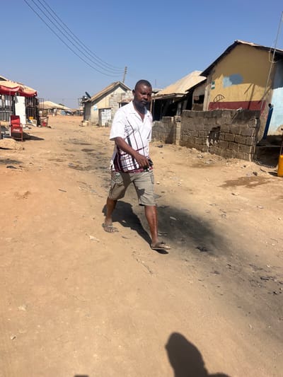 An adult male with dark skin and a beard walks on an unpaved, reddish-brown dirt path in Dzhidu, Nigeria. He is wearing a white short-sleeved collared shirt with red and black patterns, light brown cargo shorts, and two different colored flip-flops (one brown, one grey/brown with a silver embellishment). He holds a dark object in his right hand. Long shadows are cast on the ground, indicating a bright, sunny day.
The path shows tire tracks and scattered debris. To the left, several simple structures with corrugated metal roofs are visible, along with a red Coca-Cola cooler and a red canopy. To the right, a building with a yellow facade featuring blue and red markings stands behind a partially damaged concrete block wall. A yellow plastic jerrycan rests on the far right. Utility poles and overhead power lines traverse the clear blue sky in the background.