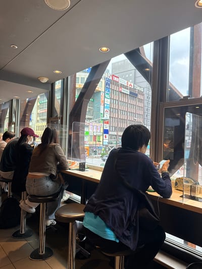 In Shinjuku, Japan, four individuals are seated at a counter facing a large window that overlooks a busy city street. The individuals are positioned from left to right: one person with their back to the camera, wearing a dark shirt, another person wearing a red baseball cap and a white mask, a third person with their back to the camera wearing a brown jacket and blue jeans, and a fourth person, also with their back to the camera, wearing a dark blue long-sleeved shirt and dark pants, holding a mobile phone. The counter is equipped with clear plastic dividers between each seating area. The window reveals a cityscape with tall buildings, advertisements, and traffic. The interior of the establishment features a dark ceiling with recessed lighting and a metallic structural element. The floor is tiled. The scene appears to be a cafe or a similar establishment providing a view of the urban environment.