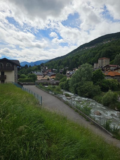 A scenic view of the Pontagna river flows through the town of Pontagna, Italy. The river is flanked by a paved path and lush green grass on the left side of the image, with a fence separating the path from the grass. On the right side of the river, trees and buildings line the hillside. The buildings are a mix of traditional European architecture, with tiled roofs and stone facades. In the background, a dense forest covers the mountainside, and the sky is filled with white, fluffy clouds. A few structures are visible on the far slope of the mountain, appearing to be houses or other residential buildings. The overall scene is peaceful and picturesque, showcasing the natural beauty of the Italian Alps.
