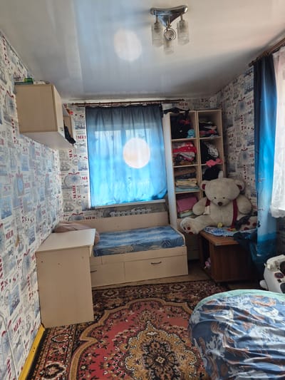 The image shows a room in an apartment located in Sokur, Russia. The room is furnished with a bed, a desk, a shelf unit, and a rug. The walls are wallpapered with a nautical theme. The bed is covered with a blue and white patterned comforter. The desk is light wood and is positioned to the left of the bed. The shelf unit is also light wood and contains clothing and other items. A large stuffed teddy bear is sitting on a wooden table to the right of the shelf unit. A window with blue curtains is visible behind the bed. The floor is covered with a red and gold patterned rug. The room appears to be a bedroom or a living area.