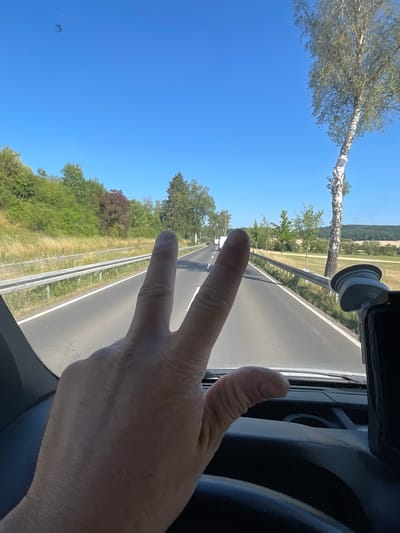 The image shows a first-person perspective from inside a vehicle, likely a car or a truck, on a sunny day. The road ahead is visible, with a metal guardrail running along the right side. The surrounding environment is rural, featuring a grassy embankment on the left and trees and fields on the right, stretching into the distance. A tall, slender birch tree with distinctive white bark stands prominently to the right of the road. The sky is a clear, bright blue. In the foreground, a human hand is held up, forming a peace sign with the index and middle fingers extended. The hand appears to be resting on the steering wheel or dashboard of the vehicle. There are other vehicles visible further down the road. The scene is set in or around Wartenberg, Germany, suggested by the location information.