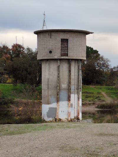 A cylindrical brick tower with a boarded-up window and a smaller hole sits amid trees and foliage. The tower's lower section has faded, painted stripes with exposed rust and faded "KEEP OFF" markings. A path and hillside rise on the right side of the frame. An antenna is perched atop the tower, and the sky is overcast.