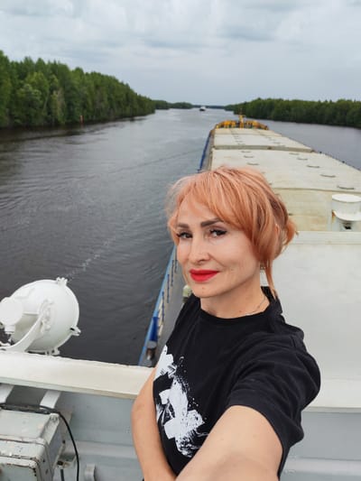 A woman with short, reddish-blonde hair and bright red lipstick smiles for a selfie on the deck of a large boat. She is wearing a black t-shirt with a white graphic design. The boat appears to be a cargo ship or barge, with a long, flat deck visible behind her. The boat is on a wide river, with lush green trees lining the banks on either side. The sky is overcast, suggesting a cloudy or possibly rainy day. In the distance, other boats can be seen on the water. The scene appears to be set in a natural, possibly rural, area given the dense forestation. The woman's expression is pleasant and self-assured. There is no visible text in the image. The location is identified as Novokemskii, Russia.