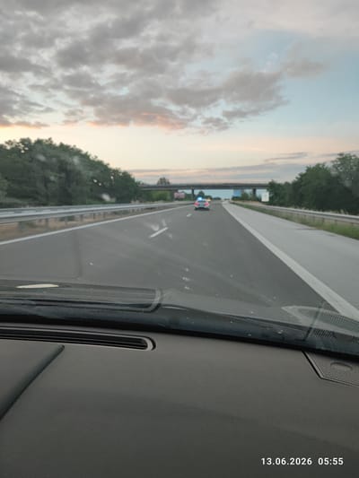 An image taken from inside a vehicle shows a highway at dusk. The sky is filled with clouds tinged with pink and orange from the setting sun. On the right side of the highway, a dense line of trees forms a barrier. In the distance, a highway overpass spans across the road, and a police car with flashing blue lights is visible on the highway ahead. The foreground of the image is dominated by the interior of the vehicle, including the dashboard and the windshield. The date and time stamp on the image reads "13.06.2026 05:55".