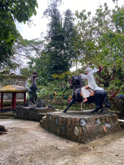 The image depicts a scene in Kecamatan Manyaran, Indonesia, located at coordinates -7.826 latitude and 110.816 longitude. Two statues are positioned on a pathway, one depicting a dinosaur and the other a person riding a horse. The statues are made of concrete. The dinosaur statue is grey and black, standing on its hind legs, facing forward. The person on the horse statue is dressed in white robes and is holding a whip. The horse statue is black, facing right. The statues are positioned on small, low platforms. The background is filled with foliage, including tall trees and smaller shrubs. The ground is dirt and gravel. The scene is located in an outdoor public space, likely a park or garden. The overall atmosphere is calm and peaceful.