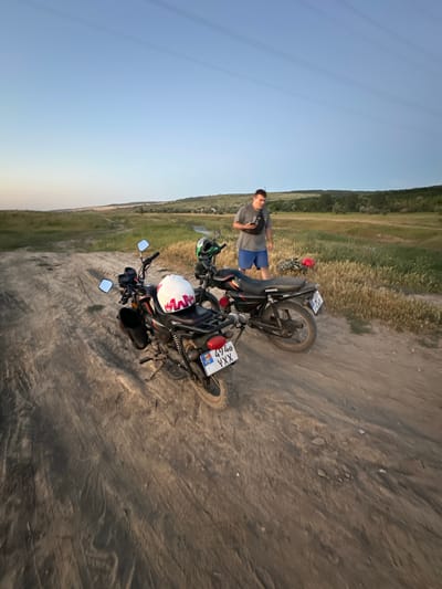 In a rural setting of Filipeni, Moldova, a young man stands next to two motorcycles on a dirt path. The scene is illuminated by the soft light of dusk, with a clear sky transitioning to a gentle gradient of blue and yellow. The ground is dusty and uneven, with tire tracks indicating recent activity.

The man, dressed in a grey t-shirt and blue shorts, has a dark fanny pack slung across his chest. He is looking towards the camera with a neutral expression. One motorcycle, primarily black with red accents, is parked upright in the foreground, its license plate clearly visible with the characters "XXX YXX 767". A white helmet with red accents sits on the seat of this motorcycle. Behind it, another motorcycle, also black, is angled on the ground, with a red helmet resting near its handlebars.

The background consists of a field of dry grass and a line of trees on a gentle slope, suggesting the edge of a rural landscape. Power lines stretch across the upper left quadrant of the image, adding a subtle human-made element to the natural scenery. The overall atmosphere is tranquil and suggests a moment of pause during an outdoor excursion. The time of day is likely early evening, given the warm light and the color of the sky.