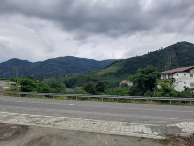 The image depicts a scenic view of a mountainous region under a cloudy sky. In the foreground, a paved road runs horizontally across the frame, bordered by a metal guardrail. Beyond the road, lush green vegetation covers the rolling hills, dotted with scattered houses and buildings. A prominent white building with a red roof stands out on the right side of the image. The landscape suggests a rural or semi-rural environment, possibly in a temperate climate. There are no people visible in the image. The scene appears to be a landscape photograph taken from a roadside perspective. This location is identified as Ayvacık, Turkey.