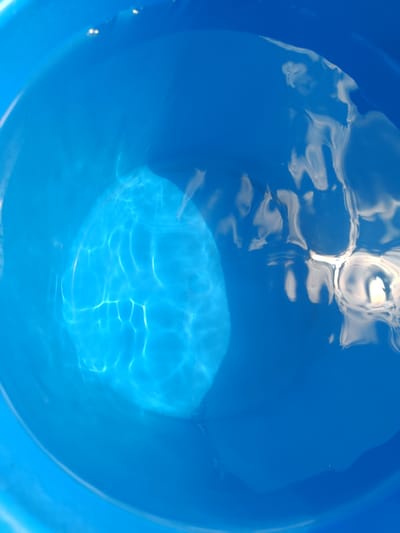 The image shows the surface of blue water, likely in a container. The water has ripples and reflections, suggesting it is outdoors or under a light source. The reflections appear to be of clouds or the sky, and possibly some man-made structures, indicating an outdoor environment. There are no people or identifiable objects within the water itself, only the patterns created by light and movement. The specific location in Zhyhalivka, Ukraine cannot be determined from the image alone.