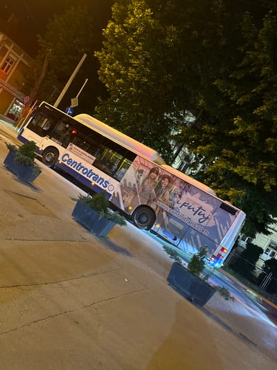 An external view of a white and blue "Centrotrans" bus parked at night on a paved street in Ilidža, Bosnia and Herzegovina. The bus features advertisements on its side, including images of people and the website "www.cent rotrans.com". The street is illuminated by streetlights, casting reflections on the wet pavement. There are planters with greenery along the sidewalk. In the background, trees and buildings are visible.