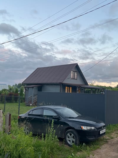 A black sedan is parked on a dirt and grass area in front of a grey wooden house with a dark brown roof in the village of Klyubushnevo, Russia. The house has windows and lights on, and there is a covered porch with string lights. A dark grey metal fence runs along the front of the property. The sky is cloudy and overcast, with power lines visible overhead. The car has a license plate that reads "Y798MA 44". The area is surrounded by greenery and trees.