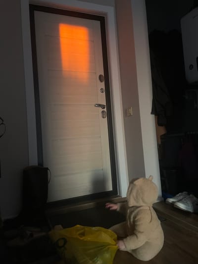 A low-angle shot shows a light-colored, paneled door with a black frame. Sunlight illuminates the upper left portion of the door. A child in a bear costume stands to the right, holding a yellow plastic bag. A dark bag sits on the floor near the door. The location is Средний Постол, Russia.