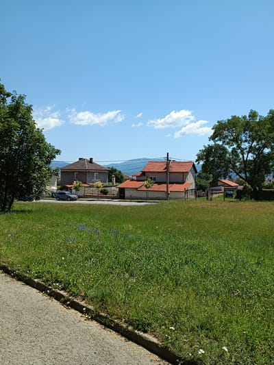 The image depicts a suburban scene in Zlatitsa, Bulgaria, on a clear, sunny day with a few scattered clouds. The foreground is dominated by a large expanse of lush green grass, dotted with small wildflowers. A paved road runs along the left edge of the frame, bordered by a concrete curb and more grass.

In the mid-ground, several houses with red-tiled roofs are visible. The house on the left is a two-story structure with a grey facade and brown trim. A dark-colored car is parked in front of it. Adjacent to this house, a fence with a wrought-iron design separates the property from the street. Further to the right, another house with a prominent red roof and lighter colored walls is situated. A black metal fence encloses this property. A tall, mature tree with dense green foliage stands to the right of this house, partially obscuring a smaller building behind it. In the distance, rolling hills and mountains form the backdrop under a bright blue sky. The overall impression is of a peaceful, residential area.