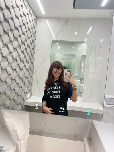 The image depicts a female individual taking a mirror selfie inside what appears to be a modern public restroom at an airport in Israel. The individual, framed from the waist up, has shoulder-length dark hair with lighter, possibly auburn, ends and bangs, and wears black-framed glasses. She is shown sticking her tongue out. Her attire consists of a black t-shirt with white text, reversed in the mirror, reading "I like more people." She wears multiple rings on both hands and numerous metallic bracelets on both wrists. Her left hand holds a light-colored smartphone, equipped with multiple camera lenses, pointed towards the mirror for the reflection.
The immediate environment features a large, rectangular mirror as the central element. Below the mirror is a white, flat counter, beneath which a white sink basin is partially visible. Integrated into the bottom edge of the mirror are illuminated icons for "AIR," "WATER," and "SOAP." The wall to the left of the individual is covered with light grey, three-dimensional hexagonal geometric panels. The wall to the right and reflected walls behind the individual are tiled with white marble-patterned material. The ceiling is white and features recessed linear LED lighting fixtures. A white double electrical outlet with two plugs is visible on the far right wall. A white paper towel dispenser is partially visible on the lower left, beneath the counter. Within the main mirror, further reflections of the room suggest additional mirrors, creating a perception of depth.