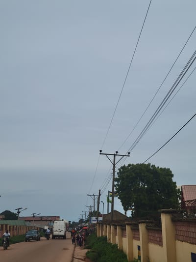 The image depicts a street scene in Tangu, Nigeria, likely during daytime given the ambient light. The sky is overcast, suggesting cloudy weather or early morning/late afternoon conditions. The photograph is oriented vertically, so the scene appears sideways.

Dominating the upper portion of the image are multiple power lines stretching across the frame, supported by wooden utility poles. A large, lush green tree stands prominently on the right side, its branches reaching towards the sky. Adjacent to the tree is a building with a light-colored facade, featuring brick accents and some windows.

Further down the street, a variety of objects and activity are visible. A white van is parked on the right side of the road, with other vehicles and motorcycles further in the distance. Several people are present, some on motorcycles and others walking. The road itself appears to be paved with a reddish-brown surface. Buildings line both sides of the street, with more greenery interspersed. One building on the right has a sign with text, though it is partially obscured and difficult to read. Another sign is visible further down the street that reads "NIGERIA". The overall atmosphere seems to be that of a busy street in a town or city.