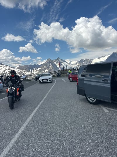 The image shows a mountain landscape at a high altitude, likely part of the Großglockner High Alpine Road, near Fusch an der Großglocknerstraße, Austria. The scene is a paved road or parking area with several vehicles. In the foreground, a person wearing a black and orange motorcycle suit and a red and black helmet rides an orange and black motorcycle. Behind the motorcycle, a white Audi sedan with a European license plate is parked. To the right of the Audi, a red car is visible, and further to the right, a dark gray van with its passenger door open is parked. In the background, snow-capped mountains rise against a blue sky with white cumulus clouds. A wooden fence runs along the edge of the road in the mid-ground, and some information boards are visible behind it. There are other motorcycles and people visible in the mid-ground. The overall scene depicts a scenic stop along a mountain road.