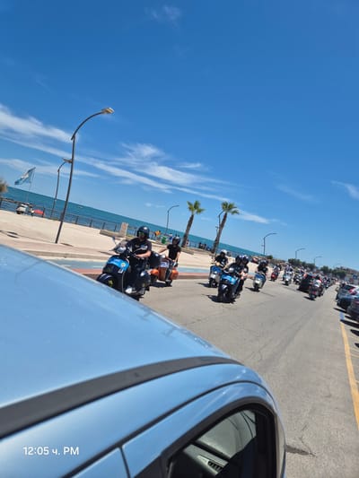 The image shows a motorcade of Vespas traveling along a coastal road. The sky is clear blue with scattered white clouds. The road is paved and bordered by a sidewalk and a body of water, likely the Adriatic Sea. On the sidewalk, there are palm trees, streetlights, and a flag. The Vespas are mostly black and white, with a few orange ones. The riders are wearing helmets and casual clothing. The perspective is from a vehicle moving alongside the Vespas, with the hood of the vehicle visible in the foreground. The timestamp "12:05 ч. PM" is visible in the lower left corner. This scene appears to be in Bisceglie, Italy, given the presence of coastal scenery and the Italian flag.