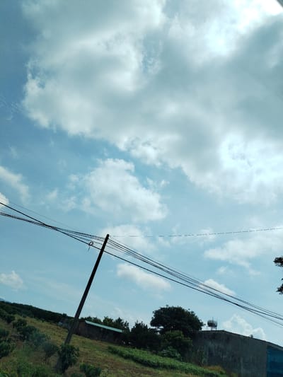The image depicts a daytime scene in Pờ Y, Vietnam. The sky is predominantly blue with scattered white clouds. A utility pole is visible in the midground, supporting multiple black power lines that stretch across the frame. The foreground features a sloping hillside covered in grass and vegetation, including trees and shrubs. A concrete wall and a water tank are visible on the crest of the hill. The light suggests it is during the day.