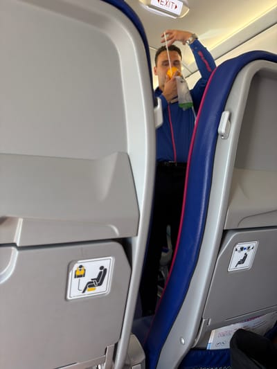 A man in a blue shirt and black pants is holding an oxygen mask in front of his face inside an airplane cabin. He appears to be demonstrating how to use the mask, with the mask hanging from a white string. The cabin interior is visible, with gray seats and blue and pink accents on the seatbacks. An "EXIT" sign is illuminated above the man. Below the man's left hand is a safety card with an icon of a person in a life vest. Another safety card is visible on the seat in front of him. The image is a medium shot, taken from a slightly low angle. The scene is forensic, matter-of-fact, and describes the physical elements of the image without interpretation. The location of Donja Cijevna, Montenegro, is not depicted or suggested by the image.