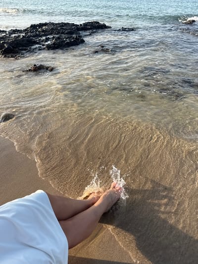 The image captures a first-person perspective of a person's legs and feet at the edge of the sea in Al Fiqait, United Arab Emirates. The person is standing in shallow, clear water that gently washes over their feet, creating splashes. They are wearing a white garment, possibly a dress or skirt, which is visible at the bottom right of the frame.
The scene is a tranquil beach setting, with golden-hued sand meeting the clear, rippling water. The water reveals the textured sandy bottom beneath. To the left, a dark, rocky shoreline or outcrop extends into the water, contrasting with the lighter tones of the sand and water. Further out, the water appears deeper and a lighter blue-grey.
The lighting suggests late afternoon or early morning, characterized by warm, golden tones on the sand and water, and the presence of elongated shadows, including the shadow of the person's upper body stretching onto the wet sand. The atmosphere appears calm and peaceful, inviting a sense of relaxation. No visible text is present in the image.