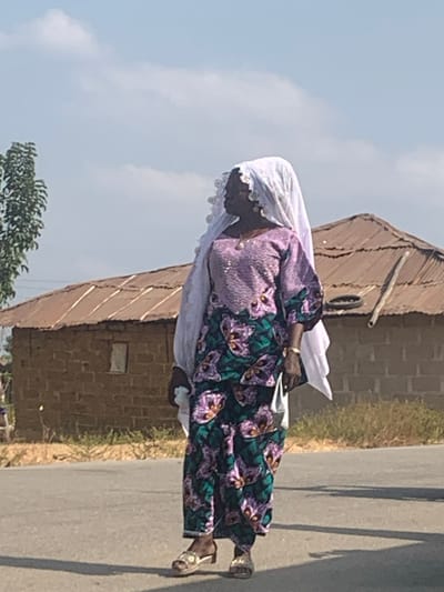 An adult female stands on a gray asphalt road under a light blue sky with sparse clouds, in the city of Nagazi, Nigeria. She is dark-complexioned, facing slightly right with her head turned to her left. She wears a white head covering with a scalloped lace edge, a light purple long-sleeved tunic with a subtle patterned texture, and dark teal trousers featuring a prominent purple and green floral design. Her footwear consists of gold-colored open-toed sandals with low heels. She accessorizes with a gold-colored necklace with a pendant and a gold-colored watch or bracelet on her left wrist, and holds a small, light-colored object in her left hand. Behind her, to the left, is a slender green tree. To her right and in the background, a single-story building with light brown brick or mud block walls and a weathered, reddish-brown corrugated metal roof is visible. A small, rectangular, light-colored window is present on the building's visible wall. A discarded dark metal ring and a wooden pole rest near the building on the right side. Sparse, dry vegetation borders the road and extends behind the structure. Bright daylight illuminates the scene, casting distinct shadows on the road surface.