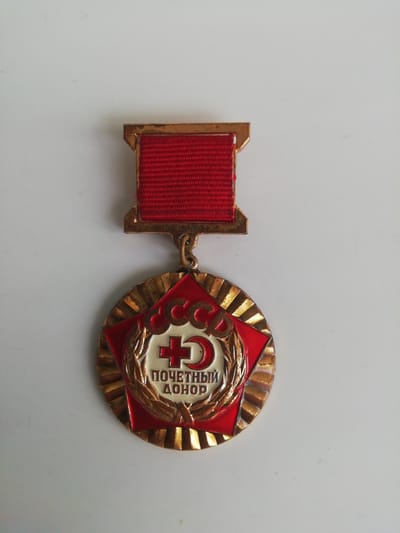 This is a close-up, eye-level shot of a Soviet-era medal displayed against a plain white background. The medal, featuring a red ribbon with a gold clasp, hangs from a gold-colored loop. The medallion itself is circular with a red star at its center, surrounded by a gold laurel wreath. Inside the star, a white emblem depicts a red cross and Cyrillic lettering.
The text on the medal reads "СССР" at the top and "ПОЧЕТНЫЙ ДОНОР" below the cross, which translates to "Honorary Donor." The overall design is detailed and ornate, with a gold, sunburst-like pattern radiating from the central emblem. The lighting is bright and even, suggesting it was taken indoors under artificial light. The white background is free of any prominent shadows or distractions, focusing all attention on the medal. There are no people, animals, or discernible environmental details present in the image. The focus is solely on the object itself.