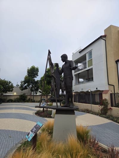 A bronze sculpture depicting a man in shorts and a t-shirt, appearing to be an artist, is set against a backdrop of a modern building and lush landscaping. The man is posed as if working on an easel, with a paintbrush or similar tool in his hand. Behind him, another, less distinct figure is visible, suggesting a possible companion or another subject.

The sculpture stands on a dark gray pedestal in an outdoor setting. The ground in front of the sculpture is paved with interlocking bricks in a patterned arrangement of light and dark gray, and beige. To the right of the paved area, ornamental grasses with a golden hue create a soft, blurred texture. Behind the sculpture and to the left, a contemporary building with white and beige facades, multiple windows, and balconies rises. Various plants, including flowering bushes with pink blossoms and darker green foliage, are interspersed around the building and along the paved pathway.

A sign with text and images is mounted on a dark pole to the right of the sculpture, near the ornamental grasses. The text on the sign is difficult to read clearly from this distance but appears to be informative, possibly detailing the sculpture or its surroundings.

The scene suggests a public space, perhaps a park, plaza, or courtyard, designed for aesthetic appreciation and possibly historical or artistic interpretation. The lighting indicates it is daytime, with an overcast sky contributing to a diffused, soft light. The overall atmosphere is calm and contemplative, with the artwork serving as a focal point in the meticulously designed landscape. The location is identified as Dana Point, USA.