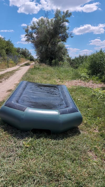 An inflatable raft, dark green with a black interior, lies on a grassy area beside a dirt path. The raft is partially filled with water. The path curves away into the distance, flanked by vegetation including tall grasses and trees. In the background, a large, leafy tree stands prominently against a bright blue sky with scattered white clouds. The scene appears to be outdoors in a natural setting, possibly a rural area or park.