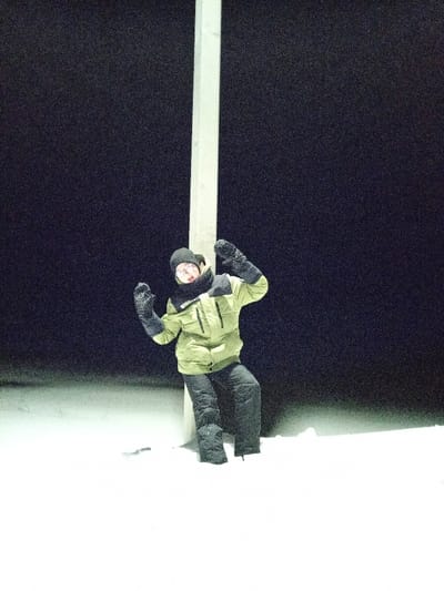 A nighttime image taken in Старый Шагирт, Russia, shows a single individual positioned in snow. The person wears a black knit beanie, glasses, a black neck gaiter, a light olive-green winter jacket with black accents, black winter pants, and black heavy mittens. They are leaning with their back against a thick, light-colored vertical post situated slightly left of the frame's center. The individual's right knee is bent, and both arms are raised at shoulder height with gloved hands angled slightly inward, in an expressive gesture. Their face is partially illuminated, showing an open-mouthed expression. The ground is covered by a uniform layer of undisturbed white snow. The background is a solid dark expanse, indicative of night, with visible granular texture. Illumination emanates from a direct frontal source, brightly highlighting the subject and the immediate snowy foreground.