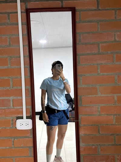 An indoor image captures an individual taking a mirror selfie. The subject is reflected in a vertical, full-length mirror with a dark reddish-brown frame, positioned against an exposed red brick wall. The individual wears a light blue athletic-style shirt with faint white patterns, blue athletic shorts, light-colored socks, and light-colored athletic shoes (possibly pink/light brown and white). Visible features include a short, dark and lighter-toned hairstyle, eyeglasses, ear piercings, and multiple tattoos on both arms and thighs. A dark smartwatch is visible on the left wrist, and a white wristband along with several bracelets are on the right wrist. A black fanny pack or belt bag is worn around the waist. The individual's face is obscured by a dark smartphone held in their right hand, positioned to capture the reflection. To the immediate left of the mirror, a white double electrical outlet (Type B) is mounted on the brick wall, with a white vertical conduit or cable cover running upwards from it. The mirror's reflection shows a white or light-colored ceiling with a visible light fixture, light-colored tiled flooring, and dark, indistinct objects in the background. The precise location within the city of Quimbaya, Colombia, cannot be ascertained from the visual evidence.