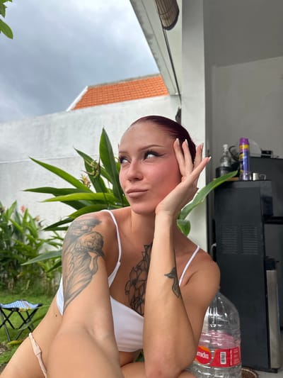 A female subject with reddish hair, a nose piercing, and a lower lip piercing is depicted in a semi-outdoor residential area within Kerobokan, Indonesia. She wears a white spaghetti strap top and light-colored bottoms. Her left hand, with light-colored fingernails, is raised to her cheek as she looks upwards and to her right. Multiple tattoos are visible, including a large design on her upper left arm, a floral pattern on her chest, and a smaller emblem on her right forearm.
The background features a white concrete wall, topped by an orange-tiled roof under a cloudy sky. To the left, a garden area with lush green tropical foliage, including plants with red and orange blossoms, is present. A blue and black collapsible stool is partially visible on the far left. To the subject's right, a dark-colored appliance, possibly a refrigerator or cabinet, is seen, with a clear bottle and a purple aerosol can on its surface. A large clear plastic water bottle, labeled "midis" and "RECYCLE ME," rests in the immediate foreground to the right of the subject.