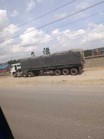 The scene is set outdoors in the city of Matungulu west ward, Kenya, under a partly cloudy sky with visible white cumulus clouds. A white-cabbed tractor unit is hitched to a multi-axle flatbed semi-trailer. The trailer's cargo is entirely covered by a dark grey tarp, secured with visible ropes or straps. The vehicle is stationary, positioned on a light brown dirt and gravel shoulder, immediately adjacent to a dark grey paved asphalt road. The road occupies the foreground and lower half of the image. Behind the truck and dirt shoulder, a light-colored concrete block wall runs horizontally. Further in the background, sparse green trees and several indistinct buildings, one with a blue roof and another with a reddish-brown roof, are visible. Multiple black power lines are strung horizontally across the upper portion of the sky. No individuals or active movements are depicted in the frame.