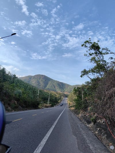 The scene captures a section of a two-lane asphalt road traversing a mountainous, vegetated landscape in Tuy Phong 4, Vietnam. The road features a solid white line on its right shoulder, a dashed white center line, and dashed yellow lane markings, curving gently as it descends into the distance. Two dark-colored sedans are observed driving away from the viewer on the far side of the road. Lush, tree-covered mountains form the backdrop, while dense green vegetation lines the left side of the road. The right side presents a drier, steeper embankment with tangled brown and green foliage, exposed soil, a visible drainage culvert, and an orange hose or cable running along its base. Utility poles with overhead lines are spaced along the left side of the road, receding into the background. A black streetlamp arm extends into the upper-left quadrant. The sky above is light blue, dotted with scattered white cumulus clouds. A blurred blue element, consistent with part of a helmet or vehicle body, occupies the extreme bottom-left foreground. No discernible people are visible.