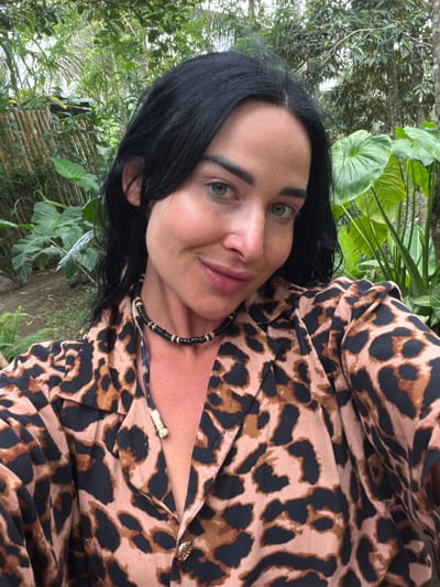 A female subject is centered in a chest-up, selfie-style portrait. She has long, dark, damp-appearing hair and blue-grey eyes, with freckles visible on her face. Her lips show a subtle smile. She wears a leopard-print collared shirt, open at the top, revealing a beaded necklace of black and tan segments around her neck. The background features dense, tropical foliage including large-leafed plants, various trees, and a partial bamboo structure on the left. Exposed ground appears as light brown soil. The scene is situated in Tegal Suci, Indonesia.