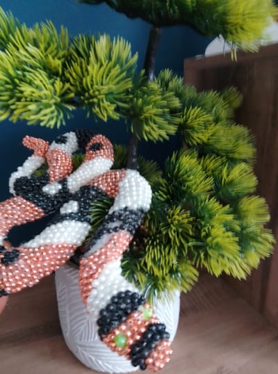 A beaded figurine resembling a panda is draped over the branches of a small artificial pine tree, which is housed in a white textured pot. The panda figurine is made of small beads in black, white, and copper colors. The artificial tree has bright green needles and is placed on a wooden surface. The background is a dark blue wall. The scene is indoors and appears to be a decorative arrangement. The location in Wedel, Germany is unknown.