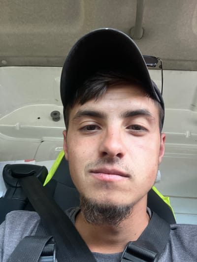 A man wearing a black baseball cap and a grey t-shirt is looking at the camera. He has a goatee and is wearing what appears to be a black shoulder strap. Part of a neon yellow safety vest is visible. The background includes a white or light grey wall with a handle and a circular fixture. The location in the city of Byala Reka, Bulgaria is not identifiable in this image.
