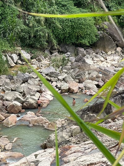 The image depicts a natural riverbed scene in Bình Lư, Vietnam. The river flows through a rocky gorge with lush green vegetation covering the slopes and surrounding rocks. Several large, gray and brown boulders are scattered throughout the waterway and along its banks. Two individuals are visible in the water. One person, wearing dark shorts, stands on a rock in the foreground, facing away from the camera. Another person is partially submerged in the water in the background. The water appears clear and greenish, with ripples indicating movement. The foreground is partially obscured by out-of-focus green foliage.