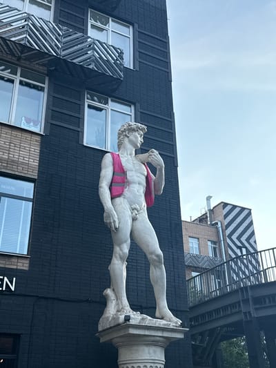 A full-body, slightly low-angle, exterior, medium shot captures a marble statue of a man, resembling Michelangelo's David, standing on a pedestal in front of a dark brick building. The statue is wearing a bright pink reflective vest over its torso. The building features several windows with white frames and blinds. To the left of the statue, a brick building section is visible, with a window displaying a sticker. Above, a section of the dark building has a modern architectural element with a patterned design. To the right of the statue, a metal walkway or bridge with railings is visible, extending across the frame. The sky is a light blue with a few faint clouds. The scene is likely located in the city of Zhabkino, Russia, as indicated by the presence of the text "МЕБЕЛЬ" (furniture in Russian) visible on a window of the dark brick building.