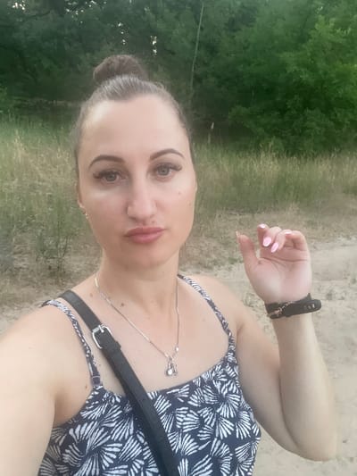 This media file features a close-up, selfie-style shot of a young woman with her hair pulled back in a bun. She is wearing a dark blue and white patterned dress and a black watch on her left wrist. A silver necklace with a small charm is visible around her neck. The woman is looking towards the camera with a neutral expression. Her left arm is bent and resting on her head, with her hand near her face.
The background is a mix of green foliage and sandy ground, suggesting an outdoor setting, possibly in a park or natural area. The lighting indicates it is daytime, with natural light illuminating the scene. The orientation of the image suggests it was taken vertically. The overall impression is a casual, personal photograph taken during an outing in Zarichne, Ukraine.