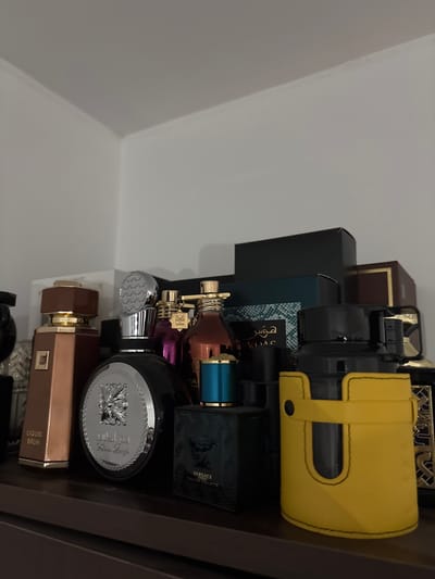 The image shows a shelf containing an assortment of perfume bottles and boxes. In the foreground, on the left, is a tall, copper-colored bottle with a gold cap. To its right is a round, black bottle with a silver, textured cap. Further to the right are several other perfume bottles of various colors and shapes, including a teal-capped bottle and a dark green bottle with "VERSACE EROS" and "EAU DE TOILETTE" visible. The background is mostly obscured by the perfume bottles, but appears to be a shelf against a white wall. The lighting is dim and suggests an indoor setting. The image is focused on the perfume collection, with no people visible. The location is specified as Smyadovo, Bulgaria.