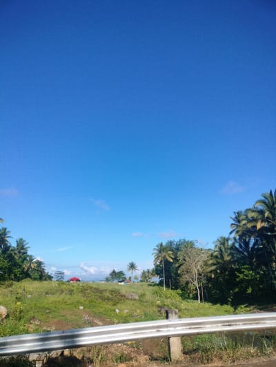 The image captures a rural landscape under a clear blue sky with sparse white clouds. The foreground is dominated by a silver metal guardrail, with dry grass and dirt visible beneath it. Beyond the guardrail, a grassy hill rises, dotted with rocks and small vegetation. A red-roofed building is situated on the top of the hill, with other structures and more greenery in the distance. Palm trees and lush foliage line the middle ground and extend into the background, suggesting a tropical environment. The overall scene depicts a serene, undeveloped area, likely on the outskirts of Kidapawan City, Philippines, characterized by its natural beauty and sparse residential presence.