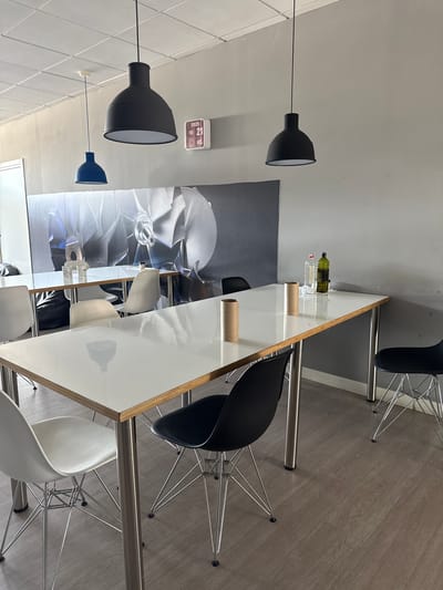 This image depicts a modern office or co-working space with a minimalist aesthetic, likely located in Getafe, Spain. The scene features two long, white-topped tables with chrome legs, arranged parallel to each other. Several chairs are placed around the tables, including white Eames-style chairs and black Eames-style chairs. The walls are painted a neutral gray, and a large, black and white mural of a turbine or fan is prominently displayed on the back wall. Three modern pendant lights hang from the ceiling, two large black ones and one smaller blue one. On one of the tables, there are two cardboard tubes, a clear plastic bottle, and a green glass bottle, suggesting recent use or preparation for an activity. A white digital clock displaying "2025 FEB 21 WED" is mounted on the wall. The flooring appears to be a light wood-look laminate. The space is well-lit, with a ceiling grid light system visible. No people are present in the image.