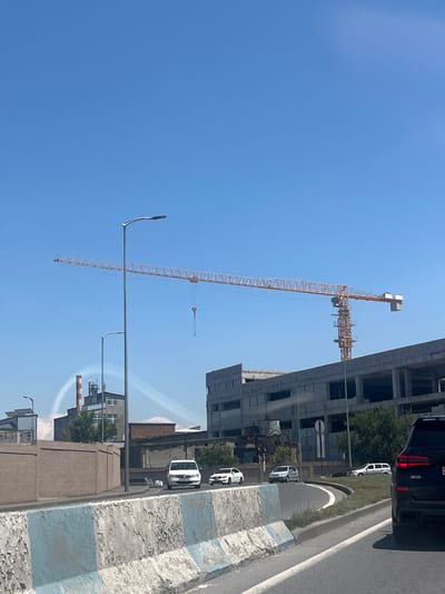 In Mkhchyan, Armenia, a large construction site is visible under a clear blue sky. A prominent yellow tower crane dominates the center of the frame, with its long boom extending horizontally across the sky. To the right of the crane, a multi-story concrete building under construction is partially visible, with exposed rebar and incomplete walls. In the background, a red and white striped smokestack emits white plumes of smoke, suggesting industrial activity. Several vehicles are present on a road in the foreground, including a white van, a white sedan, and a dark SUV. A concrete barrier with peeling blue and white paint separates the road from the construction area. The overall scene depicts ongoing development and industrial operations within the city.